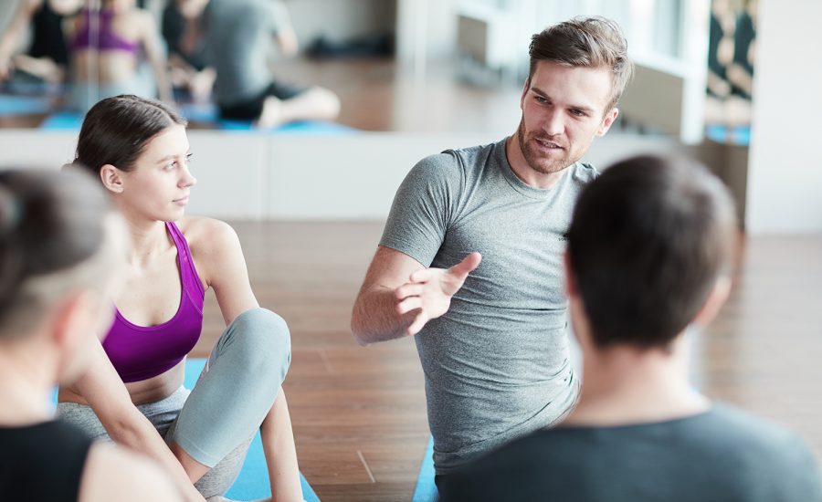 Serious pensive sporty guy with stubble sitting on floor and gesturing hand while sharing ideas for good training at yoga class